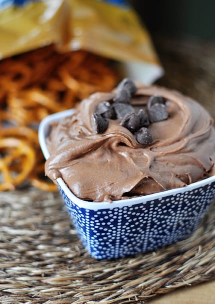 A blue ceramic bowl with chocolate brownie dip sprinkled with chocolate chips and a bag of pretzels behind it.