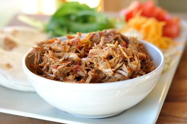 White bowl with shredded Mexican pork, on a white platter surrounded by taco toppings.