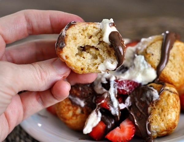 A half eaten ebelskiver in front of a plate of ebelskivers loaded with chocolate, strawberries, and whipped cream.