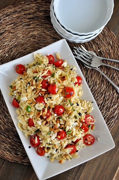 A white dish full of cooked orzo pasta, cherry tomatoes, pine nuts, and basil, with 3 forks and some white bowls on the side.