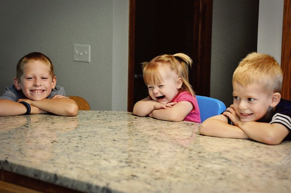 three little kids leaning their heads on a counter and smiling
