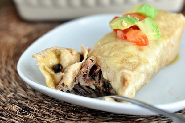 A cheesy pork burrito on a white plate with tomato and avocado dices on top.