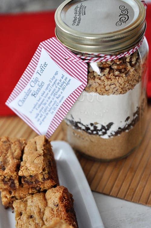 A mason jar full of layered dry ingredients to make chocolate chip toffee blondies with cooked blondies on the side.