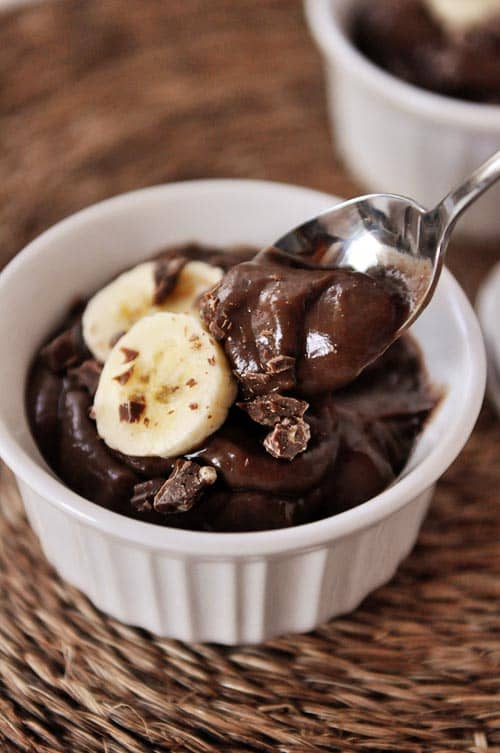 A white ramekin with dark chocolate pudding topped with banana slices and chocolate. A spoon is taking a bite out. 
