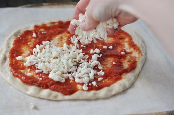 An uncooked pizza crust spread with red sauce getting cheese sprinkled on it. 