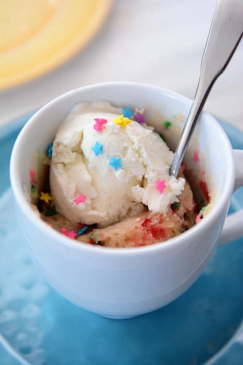 A white mug with a cooked funfetti mug cake, scoop of ice cream, sprinkles, and a spoon.