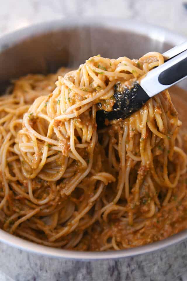 Tongs lifting up pasta and pesto sauce in stainless pot.
