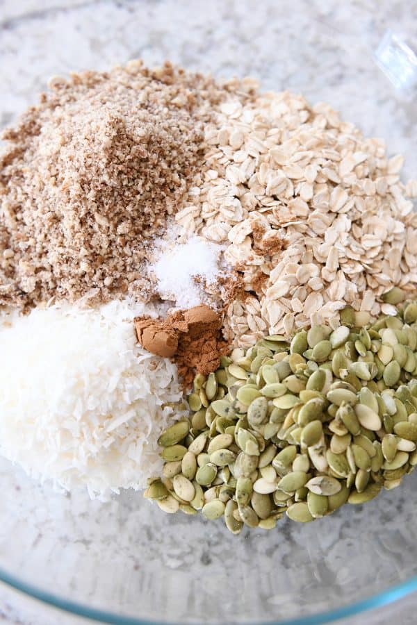 Glass bowl with ingredients for granola: coconut, pepitas, cinnamon, oats, pecans.