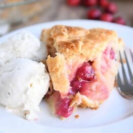 Slice of apple cranberry pie on white plate with ice cream.