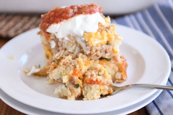 Two squares of tater tot breakfast casserole stacked on each other on white plate with sour cream and salsa and fork taking a bite