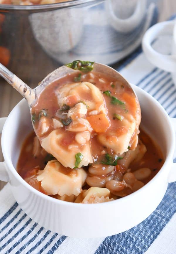 Ladle of tortellini soup going into white bowl.