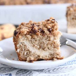 Piece of banana cream cheese coffee cake with chocolate chip streusel on white plate.