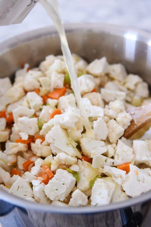 Adding broth to cauliflower, carrots, and celery for cheesy cauliflower soup.