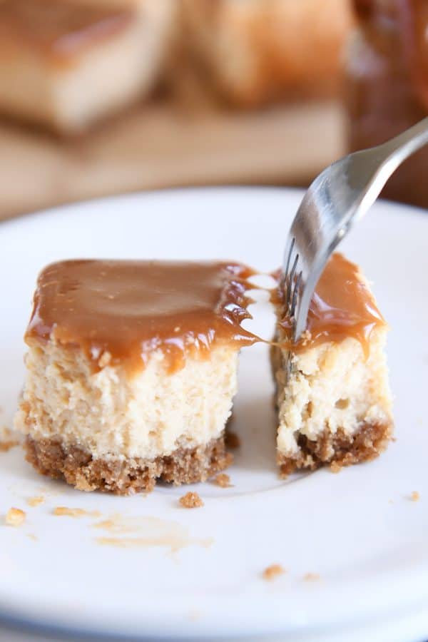 Cutting off bite of dulce de leche cheesecake square with fork on white plate.