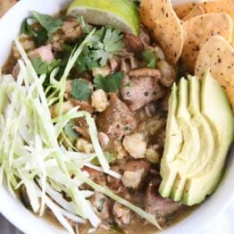 top down view of slow cooker posole in white bowl with shredded cabbage, sliced avocado, tortilla chips and lime wedge