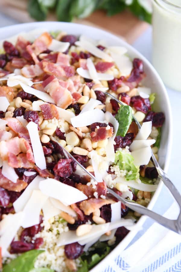White serving bowl with tongs scooping a serving of loaded couscous green salad.
