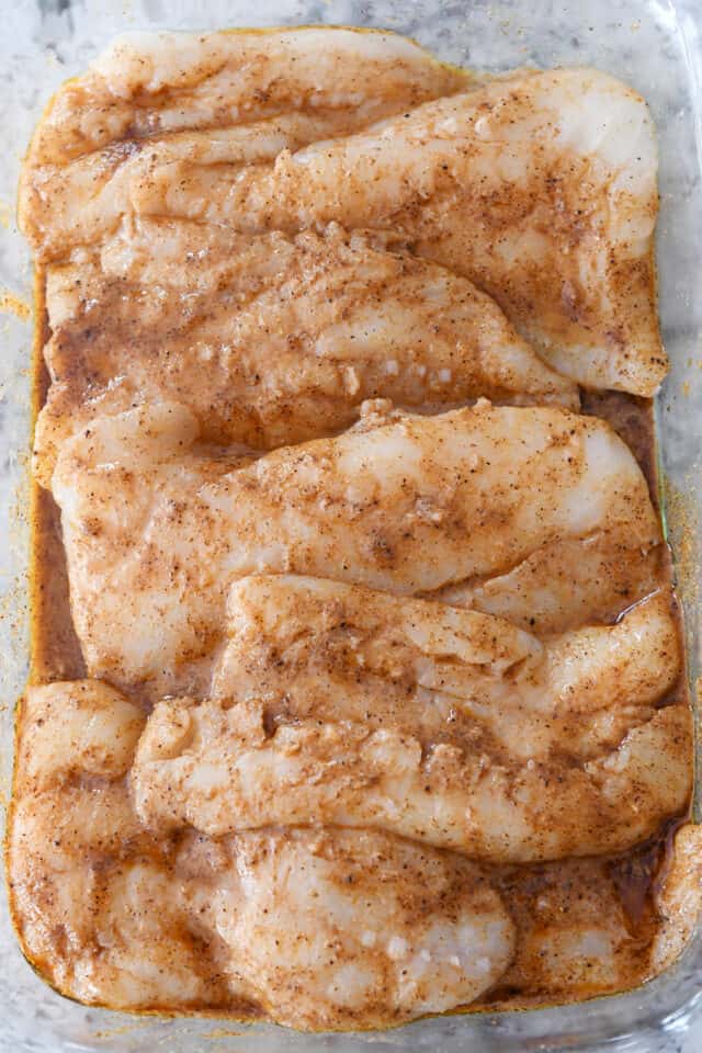 Uncooked white fish in glass baking pan with seasoning and marinade.