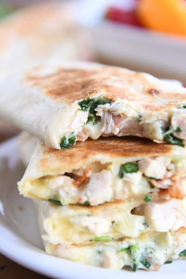 Bite taken out of crispy chicken bacon ranch wrap stacked on other wraps on white plate.