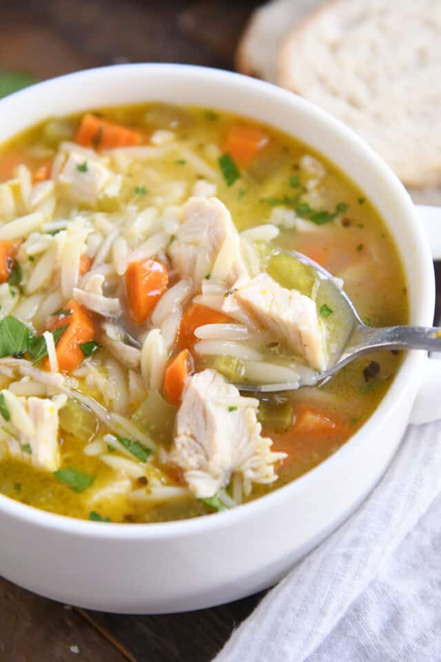 Spoon taking scoop of lemon chicken orzo soup in white bowl.