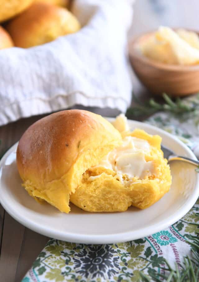 Pumpkin dinner roll in half on white plate with butter on bottom half.
