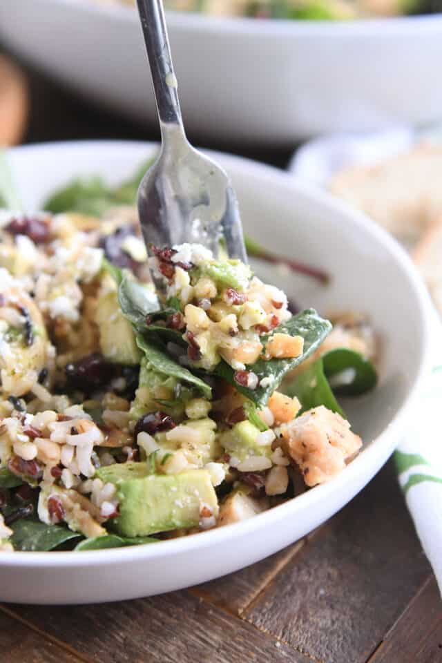 Fork scooping bite of wild and brown rice, greens, and dressing in white bowl.
