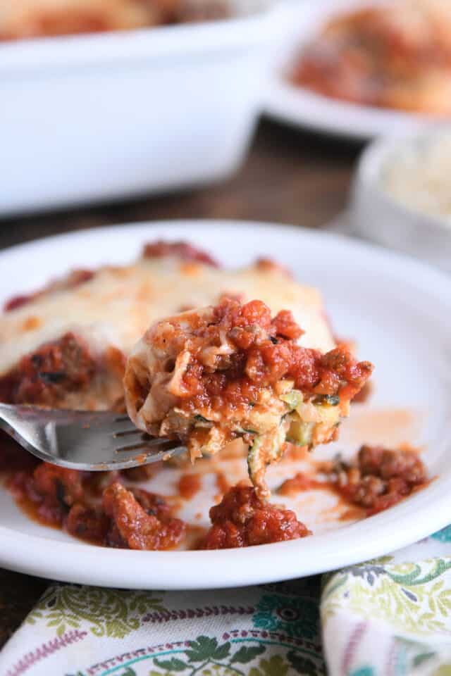 Fork scooping bite of cheesy zucchini casserole on white plate.