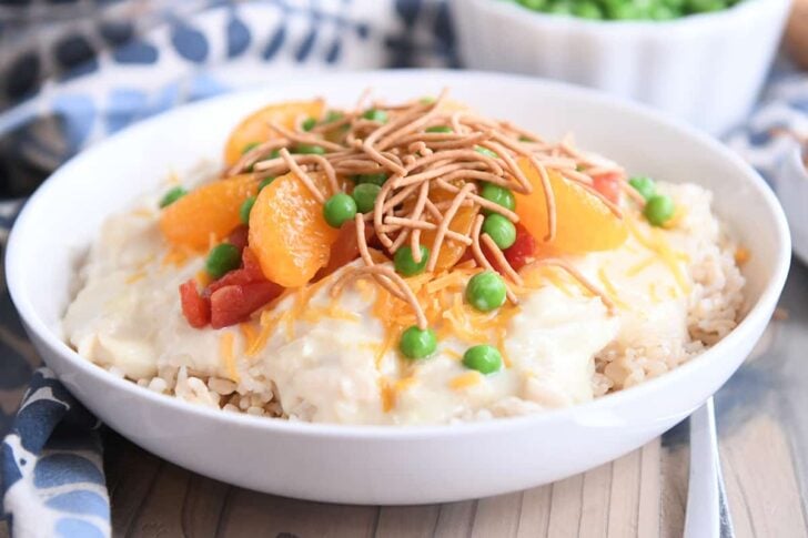White bowl with brown rice, hawaiian haystacks gravy, mandarin oranges, chow mein noodles and peas.
