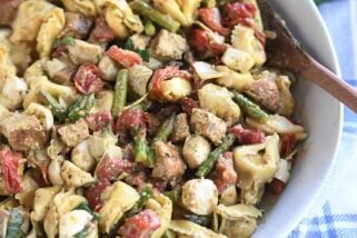 White bowl with wooden spoon on the side of tortellini salad with mozzarella, chicken, roasted red peppers and tomatoes.