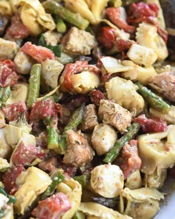 White bowl with wooden spoon on the side of tortellini salad with mozzarella, chicken, roasted red peppers and tomatoes.