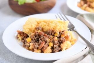 Portion of cornbread topped beef and bean casserole on white plate with fork.