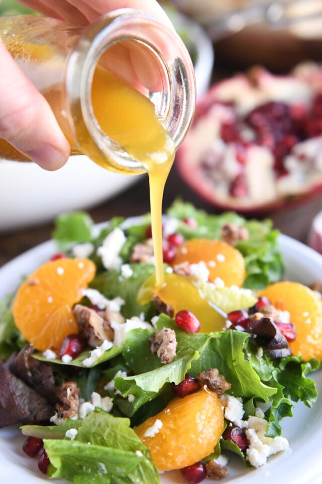 Pouring vinaigrette dressing over salad greens on white plate.