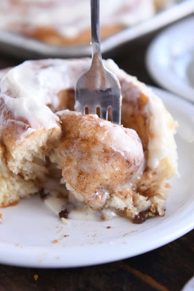 Fork digging out gooey center of cinnamon roll on white plate.