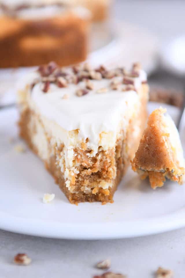 Slice of carrot cake cheesecake with bite taken out on white plate.
