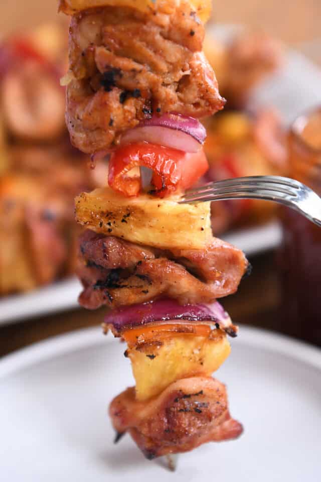 Fork pushing down pineapple on metal skewer loaded with chicken, onions, peppers.