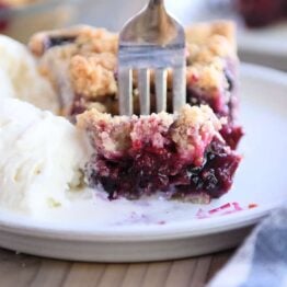 Fork taking bite of berry rhubarb pie and vanilla ice cream on gray plate.