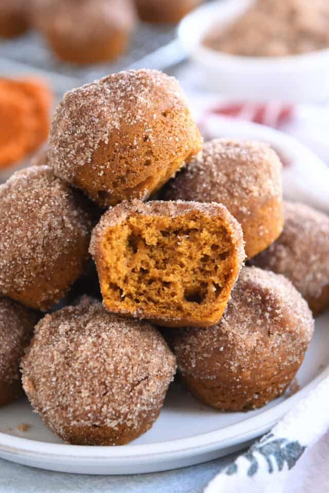 Half pumpkin mini donut muffin in stack of mini muffins on white plate.