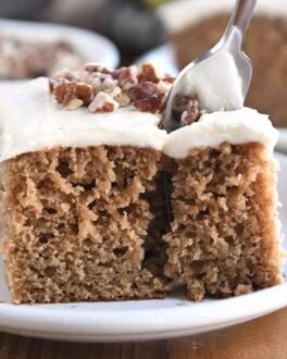Fork taking bite of frosted applesauce spice cake on white plate.