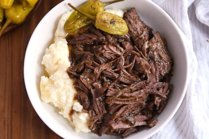 White shallow bowl with mashed potatoes, two pepperoncini, and shredded beef.