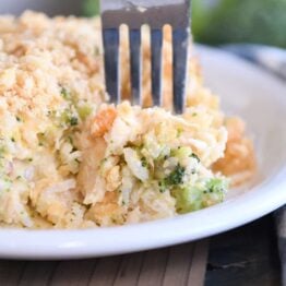 Fork scooping bite of casserole on white plate with rice, broccoli and carrots.