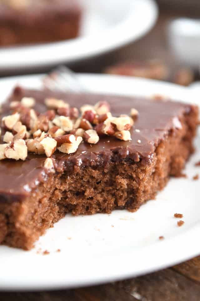 Piece removed from square of chocolate Texas sheet cake on white plate with pecans on top.