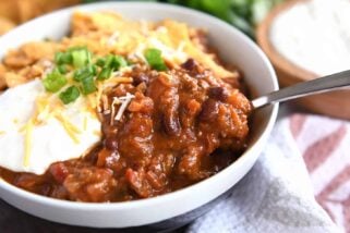 Red chili with spoonful in gray bowl with sour cream, green onions, and crushed corn chips.