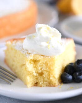 Triangle of olive oil cake on white plate with whipped cream and blueberries.