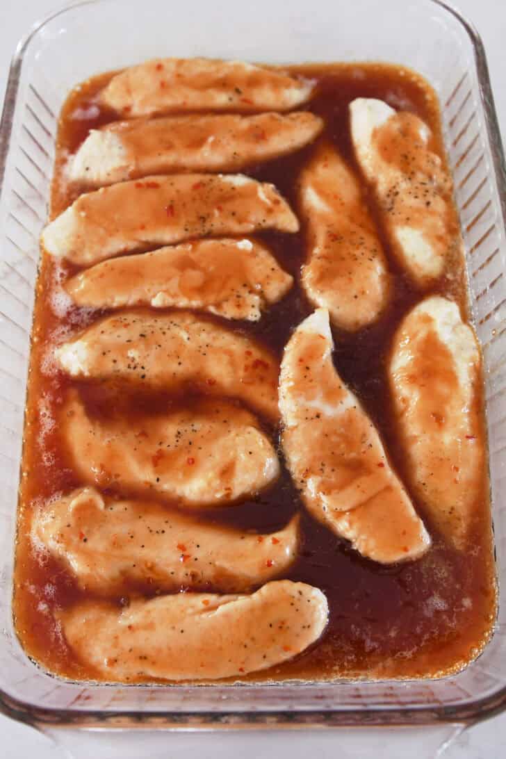 Unbaked coated chicken tenders in sauce in glass baking dish.