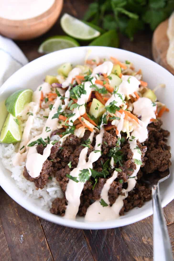 Fork in white bowl with rice, Korean beef, slaw, cucumbers, lime wedges and drizzled sauce.