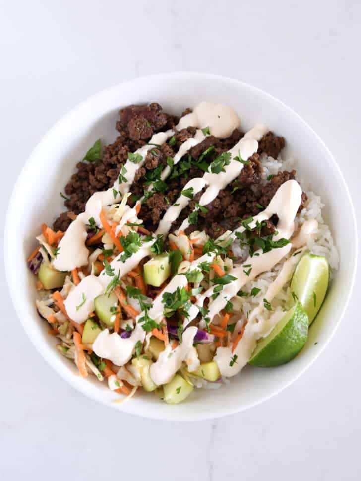 White bowl with rice, lime wedges, slaw, cucumbers, Korean beef and sauce.