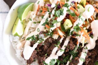 Top down view of white bowl with rice, Korean beef, slaw, cucumbers, sauce and lime wedges.