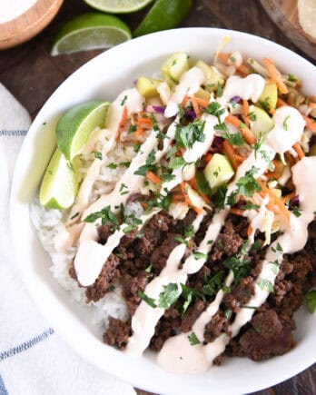 Top down view of white bowl with rice, Korean beef, slaw, cucumbers, sauce and lime wedges.