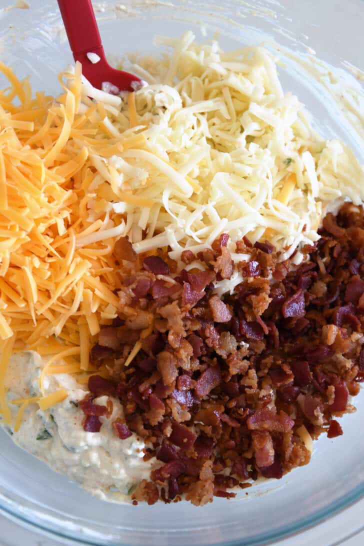 Glass bowl with creamy dip, cheddar cheese, monterey jack cheese, and chopped bacon.