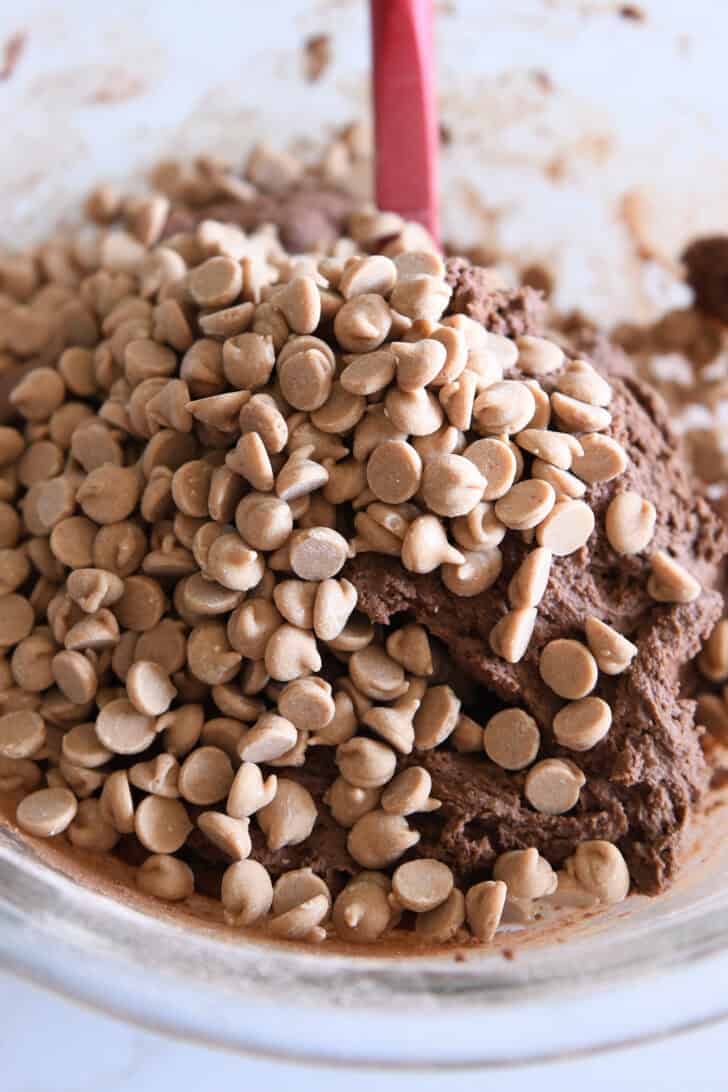 Peanut butter chips on top of mound of chocolate cookie dough in glass bowl with red spatula.