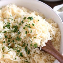 Wooden spoon scooping white rice with fresh parsley in white bowl.
